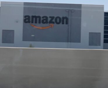 Logo de Amazon en el exterior de un centro de fulfillment con camiones de reparto y cielo despejado